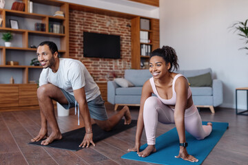 Obraz premium Smiling millennial african american man and female in sportswear doing body exercises in living room interior