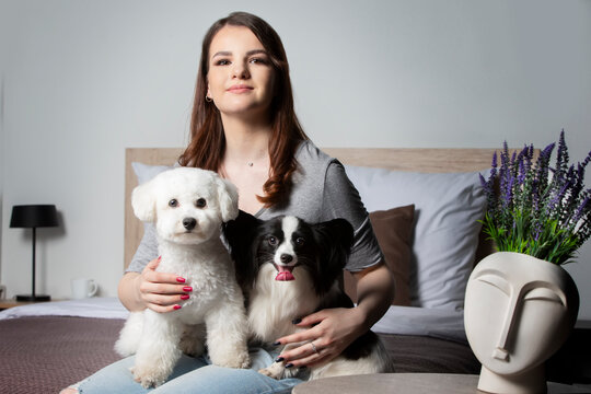 Beautiful Girl At Home With A Dog. A Hostess With A Bichon And A Papillon Sits On The Bed.