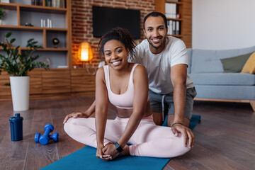 Obraz premium Cheerful millennial african american man helping to woman in sportswear doing leg stretching in living room