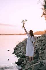 Sensual girl in white vintage dress stands with flower bouquet near the river on stones. Harmony and rest with nature. Ukrainian tradition celebration of Ivana Kupala. Quiet place.