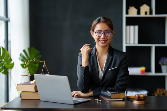 Beautiful Asian woman lawyer sitting at a table smiling happy with a laptop computer with law books hammer and scales hammer and scale.