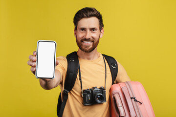 Happy man showing smartphone empty screen, wearing backpack and camera, standing with suitcase,...