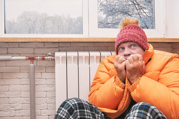 Frightened man feeling cold in hat and down jacket sitting close to radiator