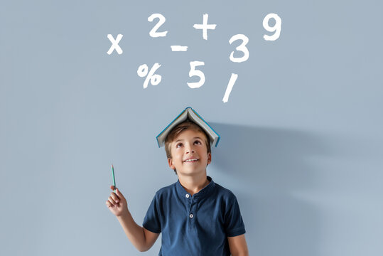 Smiling Caucasian boy with a book on his head pointing to various mathematical symbols with a pencil. Back to school, time to study subjects with numbers