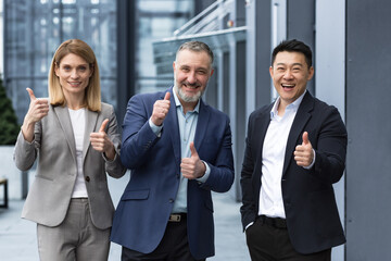 Fototapeta na wymiar Cheerful dream team, senior and experienced IT professionals in business suits smiling and happy holding thumb up and looking at camera, diverse group of project managers outside office building.