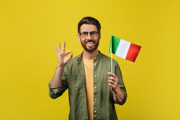 Positive man showing flag of Italy and gesturing okey, posing over yellow studio background and...