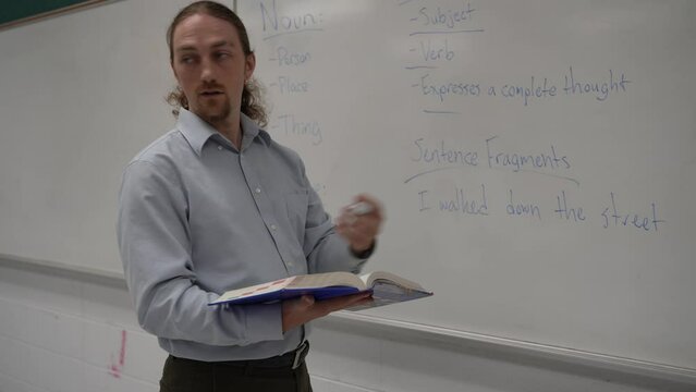 Male school teacher holding book teaching english in school classroom at a whiteboard, answering questions.