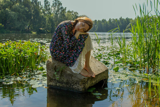 A young beautiful girl sits on a stone in the middle of a lake and dreams. Based on the painting by the artist Vasnetsov Alyonushka.