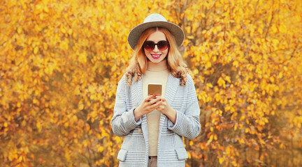 Autumn portrait stylish happy smiling woman holding phone wearing gray coat, round hat on yellow leaves background