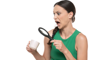 Shocked young woman looking with a magnifying glass in a cup of hot drink on a white background.