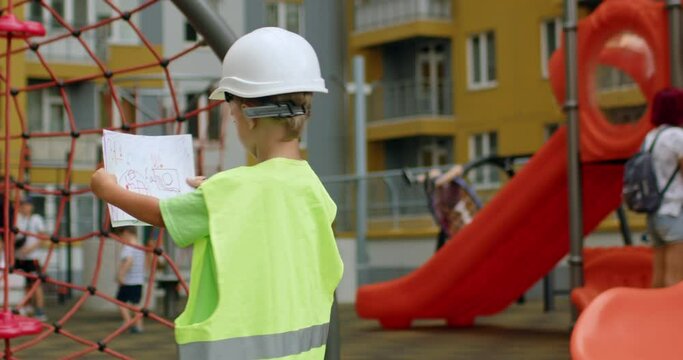 Happy preschooler boy imagines working professional builder on playground. Excited child in uniform and hardhat spins around holding papers in yard