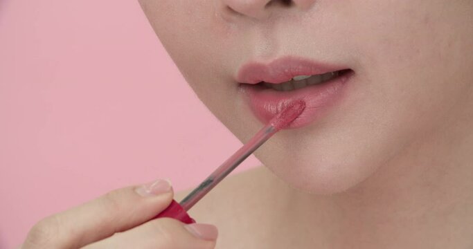 Closeup of the lower half of young pretty and beautiful Asian woman's face applying lipstick or lip gloss on her lips, smiling at camera on isolate pink background. Makeup and cosmetics concept.