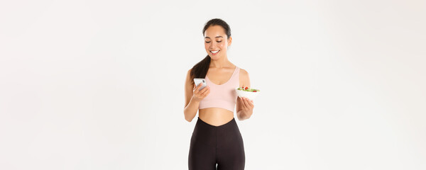 Sport, wellbeing and active lifestyle concept. Smiling cute asian girl using diet app, calories tracker application on mobile phone, contact coach to inform about food consumption, hold salad - obrazy, fototapety, plakaty