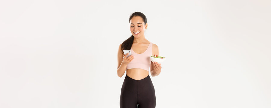 Sport, wellbeing and active lifestyle concept. Smiling cute asian girl using diet app, calories tracker application on mobile phone, contact coach to inform about food consumption, hold salad