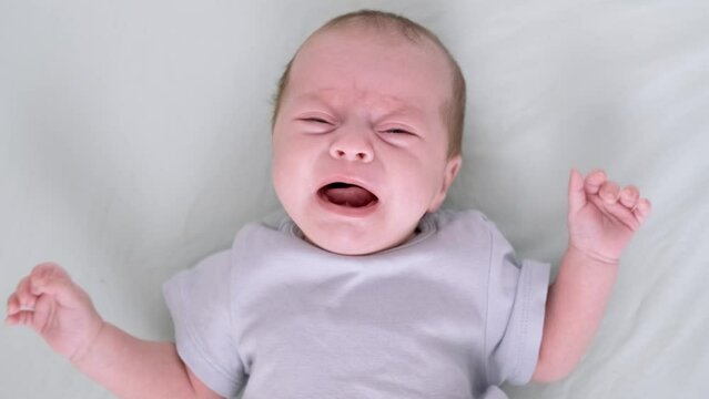 Dissatisfied, crying, upset, sad Newborn baby boy lying in nursery in crib with blue clothes on bed in bedroom at home, child crying is hungry or want to sleep