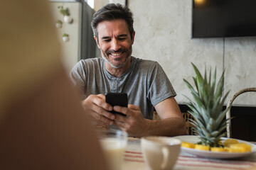 mature man with gray hair sitting in the kitchen at home smiling looking at his smartphone