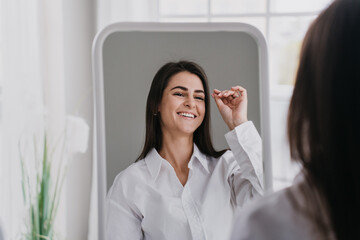 Hispanic gorgeous young woman in white shirt at home looking at mirror, toothy smiling, satisfied after cosmetology procedure, skin care, health and beauty concept. Women health and beauty.