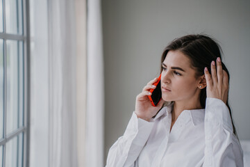 Frustrated brunette hispanic young woman talking by phone with husband, in troubles. Pretty caucasian girl at home using cell phone looking hopelessly at window. Upset housewife. Mockup sad girl.