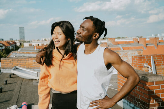 Smiling young man standing with arm around female friend looking away while standing on rooftop