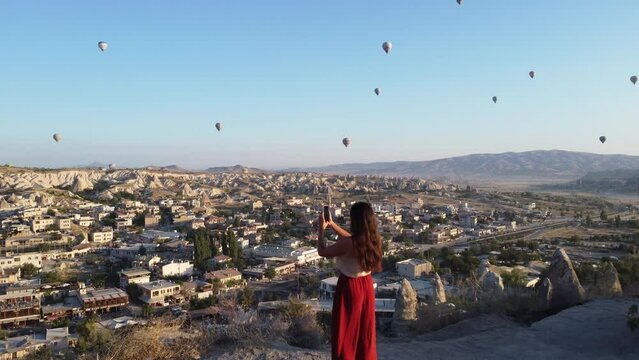 Young woman are taking photo on smartphone and enjoys the morning view on Goreme park with hot air balloons fly in Cappadocia, Turkey
