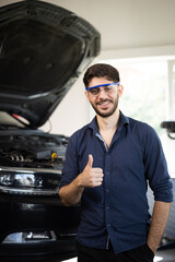 Fototapeta na wymiar Portrait of young caucasian bearded handsome man auto mechanic or manager in goggles smiling to camera and giving thumb up in garage. Cheerful guy standing at cars service