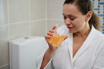 Multi-ethnic pretty woman in white bathrobe, drinking water with effervescent painkiller pills, according to physician's prescription. Woman suffering head ache. Migraine and fever, pain relievers.