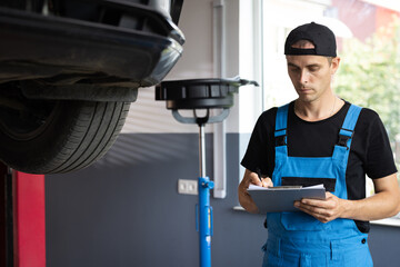 Mechanic inspects the car undercarriage way and makes a note on his inspection sheet. Car service employe inspect car. Automobile service, car mechanic. Modern workshop.