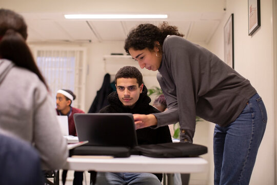 Teacher leaning on desk assisting student using laptop in classroom