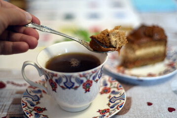 In hand, a teaspoon with an appetizing caramel cake against the background of a tea pair with tea or coffee and a saucer with a bokeh cake.