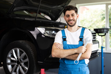 Young caucasian bearded man in blue coveralls holds spanner, smiling and looking into camera. Male car mechanic in spacious repair shop