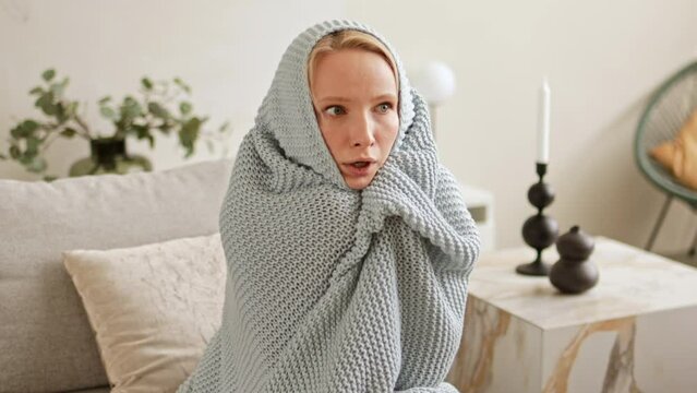 Woman is freezing and shivering from the cold, hiding herself in a warm blanket knitted plaid have to problems with central heating and heat outage at home in winter.