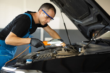 The mechanic in blue overalls and safety glasses inspects the car while working with led lamp. The work of a car mechanic in a modern workshop, car service.
