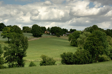 Rolling countryside with meadows, houses and trees under a cloudy sky.