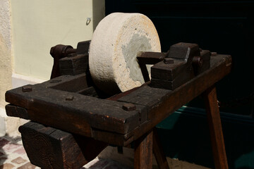 old style round grindstone wheel. knife sharpening tool set in wood frame. vintage equipment. rusty metal axel. stucco wall background. decorative piece at street storefront. rusty steel spindle. 