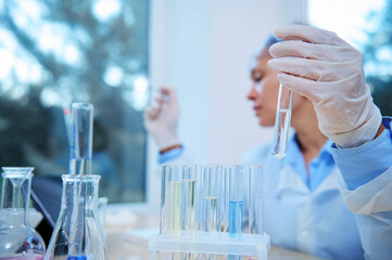 Selective focus on test tube with reagent in gloved hand of blurred woman scientist chemist experimenting chemicals, sitting at table with lots test tubes and graduated labware in research laboratory