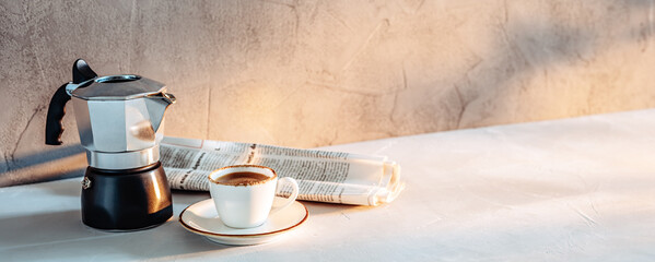 A cup of espresso, a coffee maker and a newspaper in the morning sunlight