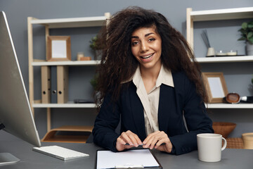 Happy friendly tanned adorable curly Latin businesswoman in jacket speak with candidate smile at camera in office. Copy space Mockup Banner. Corporation leader lady uses modern computer in work