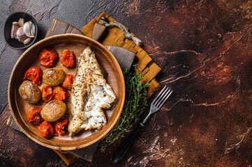 Baked halibut fish with roasted tomato and potato in wooden plate. Dark background. Top view. Copy...