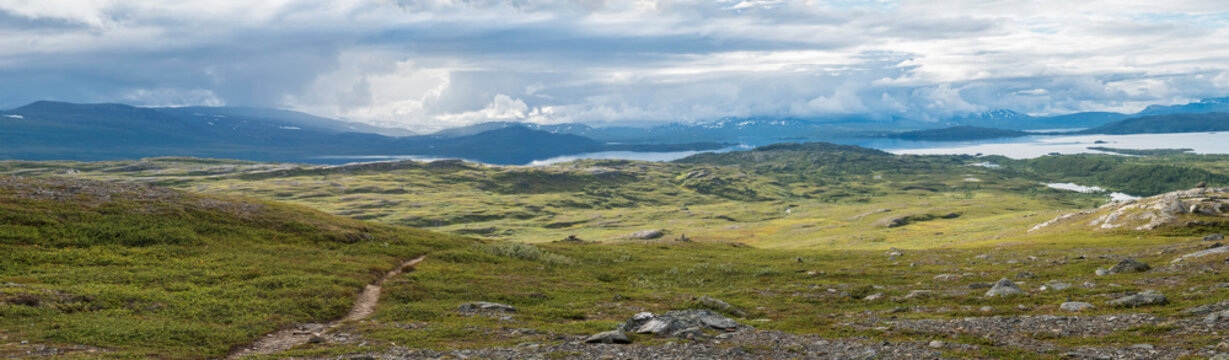 Panoramic landscape with beautiful Virihaure lake, snow capped mountain, birch trees and footpath of padjelantaleden hiking trail. Sweden, Lapland wild nature. Summer cloudy day