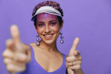 Portrait of a sporty fashion woman posing smiling with teeth and pointing a finger at the camera in a purple yoga tracksuit and a transparent cap on a purple monochrome background