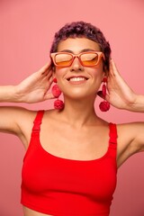 Young athletic woman with a short haircut and purple hair in a red top and pink yoga leggings in sunglasses with an athletic figure smiles and dances on a pink background