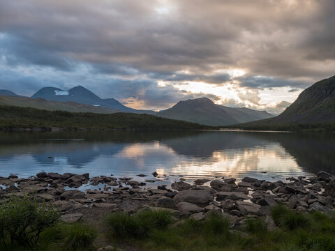 Sunset at rocky grassy shore of Tarra river. Tarraatno with green hills, birch forest and mountians at Padjelantaleden hiking trail. Summer cloudy evening with dramatic clouds at Lapland, Sweden