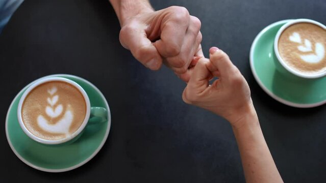 Top view coffee table with male and female hands gesturing pinky swear. Close-up unrecognizable Caucasian loving man and woman holding hands sitting indoors in cafe dating. Love and unity