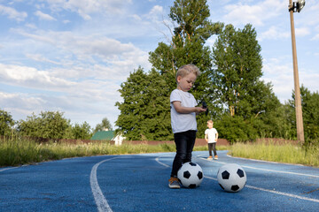 Twin brothers are playing on a walk with soccer balls.The twins are dressed the same.Children are passionate about the game.Children and football outside in sunny weather.A child with soccer balls
