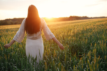 Woman walking in wheat field in warm sunset light. Stylish young female in rustic dress holding wildflowers in hands and relaxing in evening summer countryside. Tranquil atmospheric moment