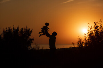 Silhouette of a man and a child against the sunset.Dad raises his son against the orange sky.People are having fun at sunset.The silhouette of a happy family standing against the sunset
