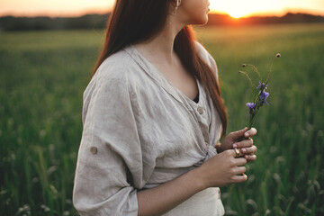 Woman holding wildflowers in wheat field in warm sunset light. Tranquil atmospheric moment. Close up of young female in rustic dress with flowers in hands in evening summer countryside.