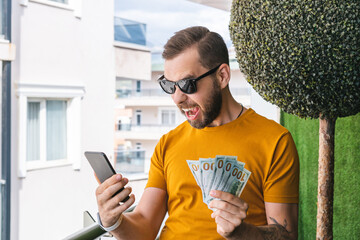 Euphoric soccer fan is watching soccer play live broadcast online on his mobile phone, betting on his favourite team and celebrating money win - 527432639
