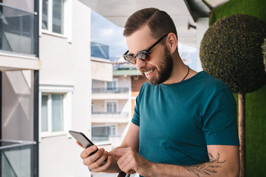 Casually dressed male travel blogger sharing new post while standing at the terrace in hotel room with the beach on the background - Powered by Adobe