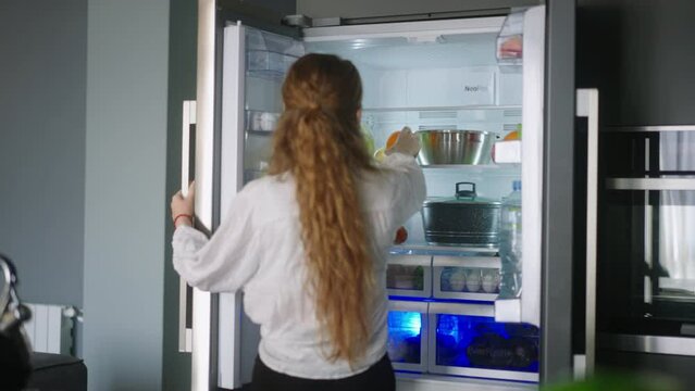 Young woman taking an orange from fridge throwing it up. Girl getting a fruit from refrigerator. Female having something healthy for snack. Caucasian woman is about to cut orange. Girl cutting orange
